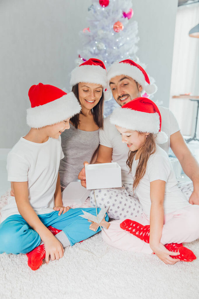 famille ouvrant leurs cadeaux ensemble le matin de Noël. ils sont tous assis par terre dans leur salon, devant l'arbre
 - Photo, image