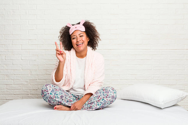 Νεαρή Αφρικάνα Αμερικανίδα στο κρεβάτι φορώντας πιτζάμα που δείχνει το σύμβολο της νίκης και χαμογελώντας ευρέως. - Φωτογραφία, εικόνα