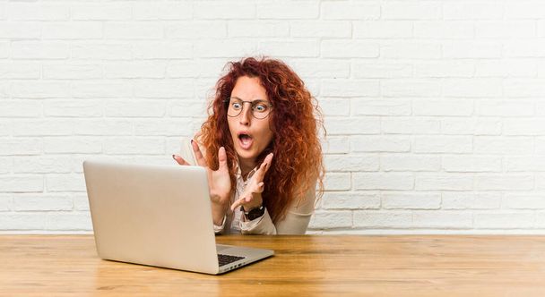 Jeune rousse bouclée femme travaillant avec son ordinateur portable crie fort, garde les yeux ouverts et les mains tendues
. - Photo, image
