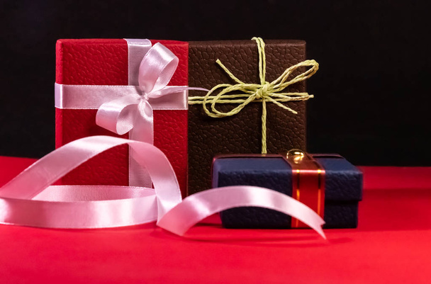 Три прикрашені коричневі, червоні та сині подарункові коробки з красивими стрічками та бантами на червоній поверхні та чорному тлі
 - Фото, зображення