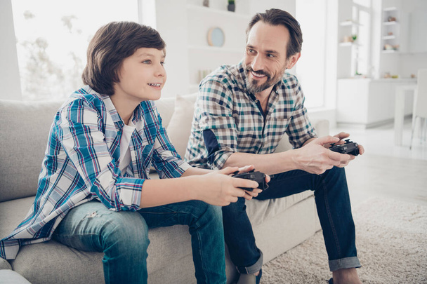 Profile side view portrait of two nice cheerful guys dad and pre-teen son sitting on sofa enjoying playing online game spending spare time at light white modern style studio interior living-room - 写真・画像