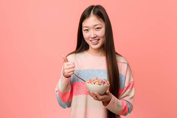 Young asian woman with a cereal bowl smiling and raising thumb up - 写真・画像