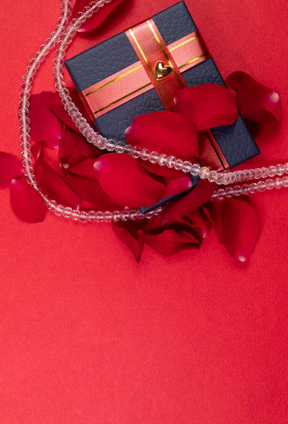 Весільний подарунковий концепт. Кришталеве намисто навколо синьої подарункової коробки з пелюстками троянд на червоному тлі зверху
 - Фото, зображення