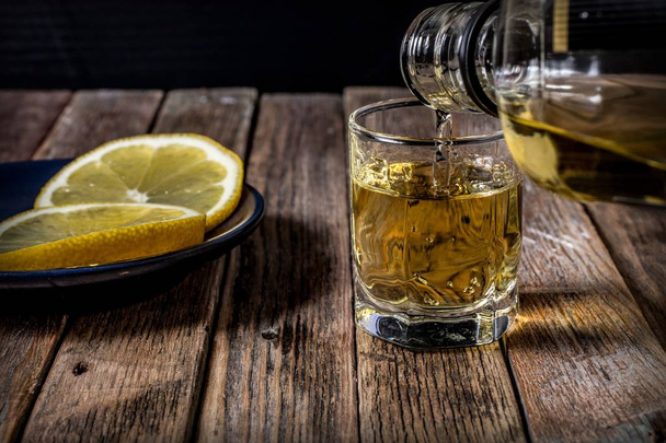 Il whisky viene versato da una bottiglia in una pila su una superficie di legno. Limone affettato
. - Foto, immagini