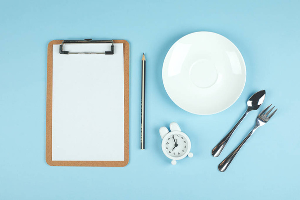 Intermittent fasting, ketogenic, diet concept. Empty plate with white alarm clock and white note notepad on blue background. Flat lay, top view, mockup - Photo, Image
