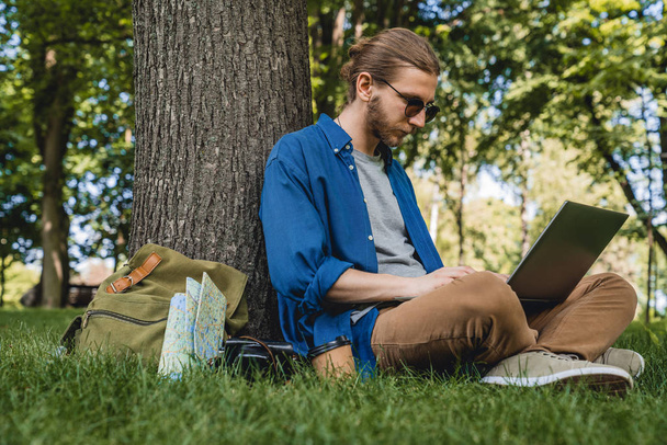 Male wearing sunglasses using laptop while sitting under a tree at park. Young man sitting on grass at park working on laptop while travelling - Foto, immagini