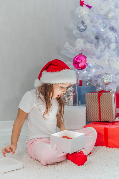 Merry Christmas celebration. Cheerful beautiful little girl sitting near the Christmas tree holding a gift box. Christmas miracles. Happy New Year. Winter, celebrating - Foto, Imagen
