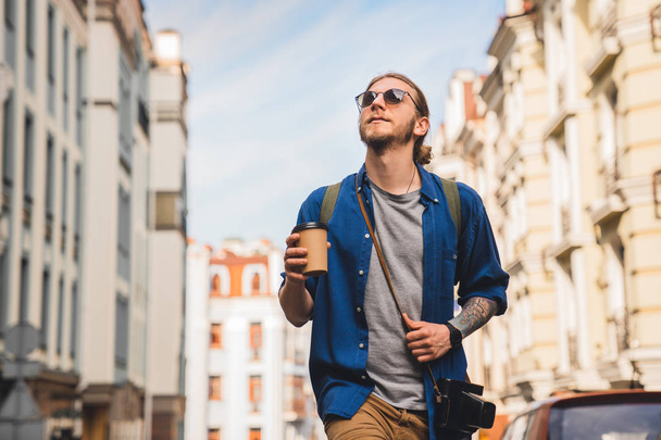 Handsome young man in casual clothing enjoying summer and wandering empty streets while walking outdoors with cup of coffee in hands - Foto, imagen