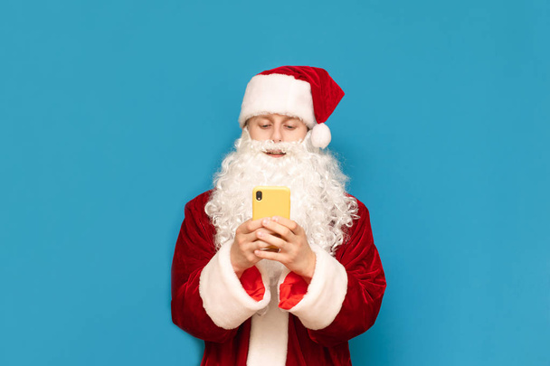 Hymyilevä nuori mies puvussa seisoo sinisellä taustalla ja käyttää älypuhelinta, eristetty. Santa teini älypuhelin eristetty sinisellä taustalla. Joulun käsite
. - Valokuva, kuva