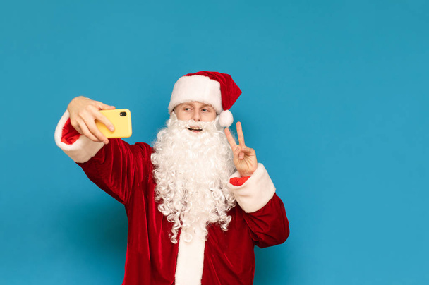 El tipo positivo en traje de Santa se toma selfie contra fondo de pared azul, mira el teléfono inteligente, sonríe y muestra gesto de paz. Joven Santa Claus tomando fotos en el smartphone. Aislado
. - Foto, Imagen