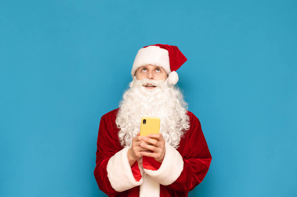 Funny young santa stands on blue background with smartphone in hands and looks up at empty space. Cheerful Santa is using a smartphone and looking up at copyspace. New Year and Christmas concept. - Foto, Imagen