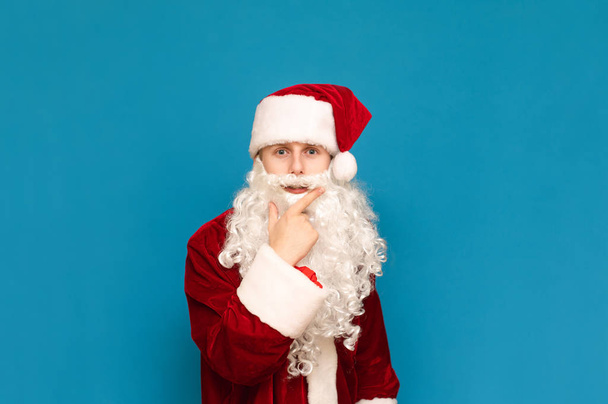 Πορτρέτο ενός αστείου σκεπτικού έφηβου σε κοστούμι Santa Claus, κοιτάζοντας έντονα στην κάμερα, απομονώνονται σε μπλε φόντο. Νεαρός Άγιος Βασίλης σε κοστούμι κοιτάζει στην κάμερα και σκέφτεται. Χριστούγεννα και το νέο έτος - Φωτογραφία, εικόνα