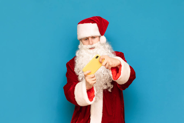 Guy in carnival costume of santa claus playing mobile games on smarphone isolated on blue background. The young site plays online games and smartphones, looks intently at the smartphone screen. - Photo, Image