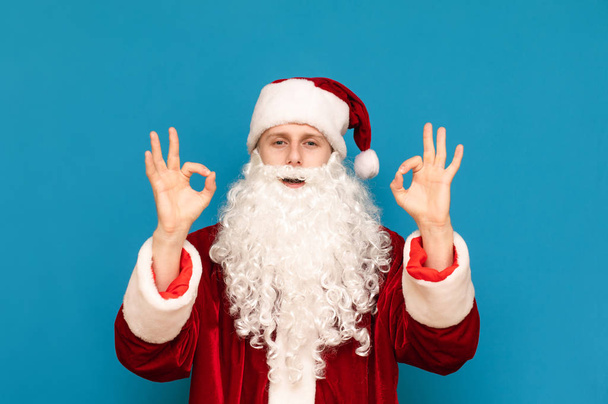 Щасливий Санта Клаус показує свої руки ОК жест, дивиться в камеру і посміхається і проти синього фону. Хлопець у костюмі Санти підняв руки і показує знак ОК пальцями - Фото, зображення