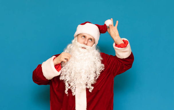 Positieve jonge Kerstman poseren op camera geïsoleerd op blauwe achtergrond. Kerel in Santa pak staan tegen de blauwe muur achtergrond en kijken naar de zijkant. Kerstconcept. - Foto, afbeelding