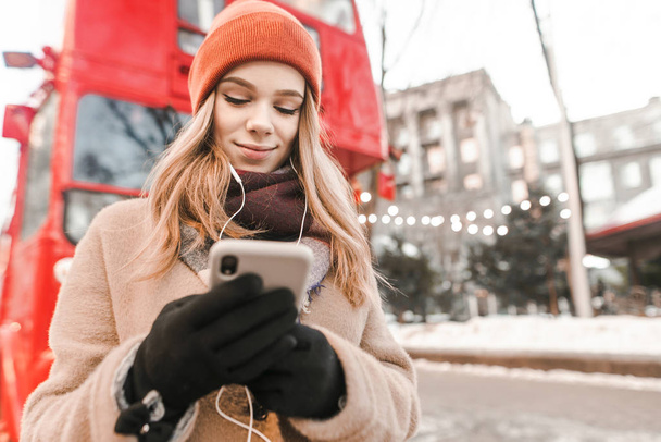暖かい服や手袋のかわいい女の子が通りに立って、ヘッドフォンで音楽を聞いて、都会の冬の背景にスマートフォンを使用して、音楽と美しい女の子の冬の散歩 - 写真・画像