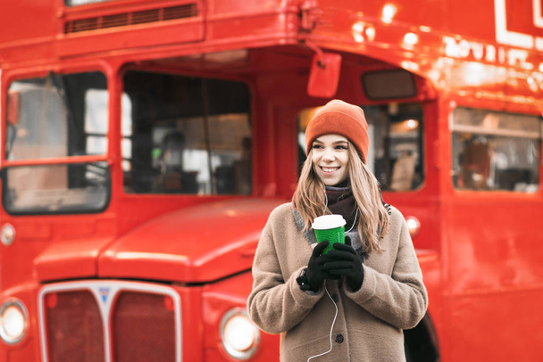Soğuk bir kış gününde yürürken sıcak giysiler içinde mutlu bir kızın portresi elinde bir fincan kahveyle kırmızı bir otobüsün arka planında duruyor ve başka tarafa bakıyor. Noel konsepti. - Fotoğraf, Görsel