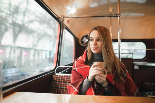 Cute girl in plaid sits in a cozy cafe with a cup of hot drink in her hand, looks out the window with a thoughtful face. Handsome lady spends time in winter season warming up in coffee shop. - 写真・画像