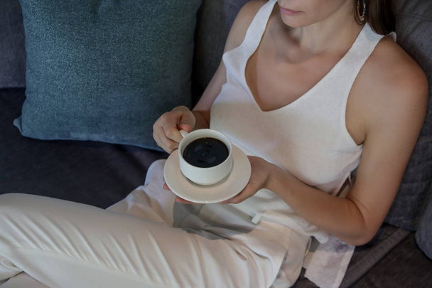 Mladá žena v bílém oblečení sedí na pohovce a mají šálek kávy, relaxaci a útulný volný čas doma - Fotografie, Obrázek