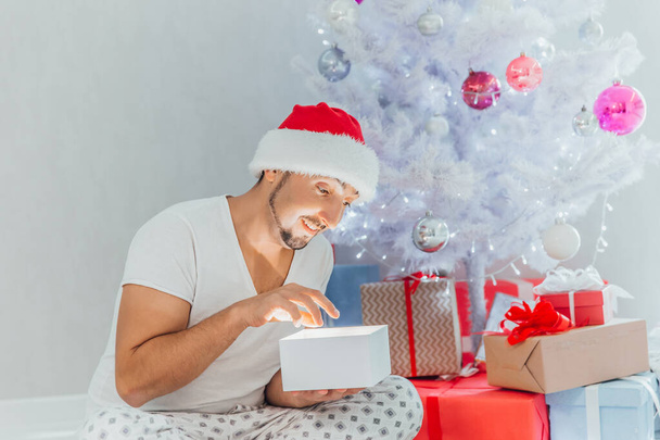 A young man in Santa hat sits under a Christmas tree and opens a gift box against the white lights in the early morning - Foto, Imagen