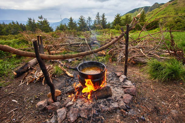 Old small kettle is heated on a bonfire on a green mountain meadow during a bad weather. - Photo, Image