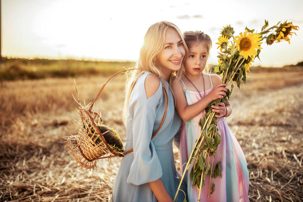 Smiling young mother walks with her little charming daughter with a bouquet of sunflowers and a basket on a warm summer evening in a field outside the city. Family walk concept - 写真・画像