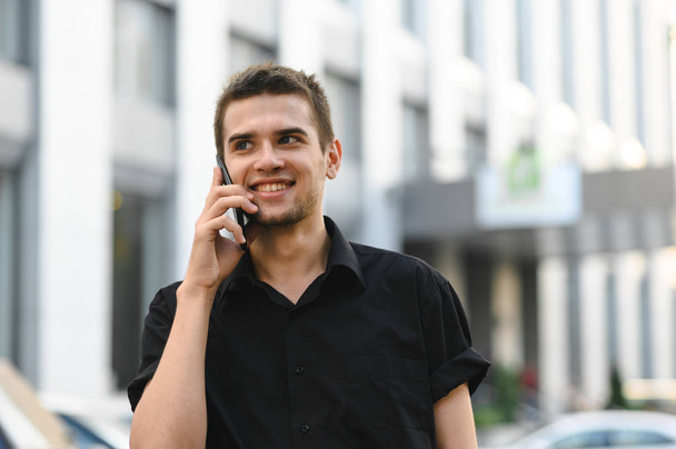 Smiling young man in black shirt talking on the phone on a bright cityscape, smiling and looking away. Stylish guy talking on the phone. Street closeup portrait. - Photo, Image