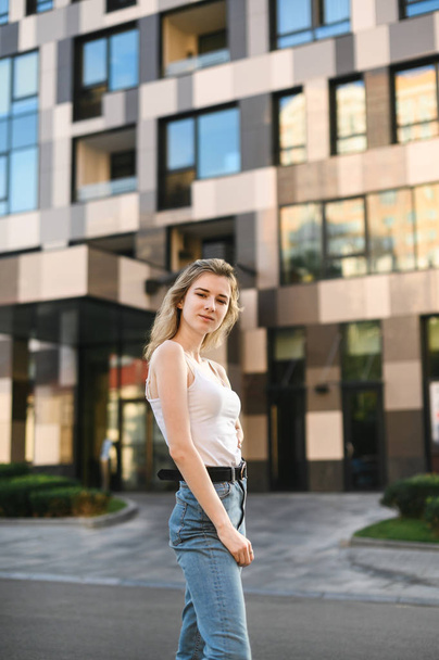 Retrato de chica atractiva en ropa casual de pie en la calle contra el fondo de un edificio moderno, mirando a la cámara, con pantalones vaqueros y una camiseta blanca.
 - Foto, imagen