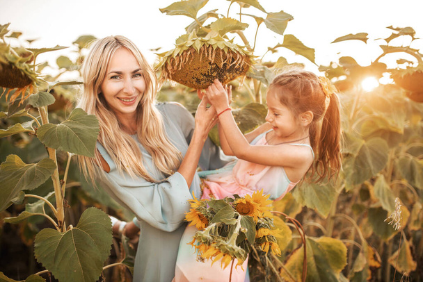 Pretty young blonde mother holds in her arms pretty little daughter with a bouquet of sunflowers while walking on the field on a warm summer evening. Nature enjoyment concept - Фото, зображення