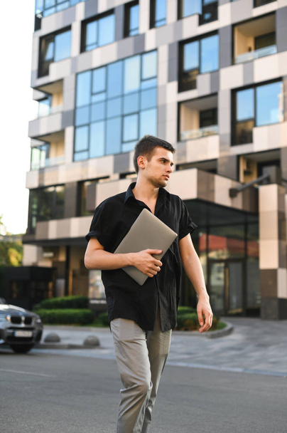 Estudiante porteador de moda en ropa casual caminando por la calle con un portátil en la mano, con una camisa negra y pantalones grises. Retrato estilo calle de un tipo con estilo
. - Foto, Imagen