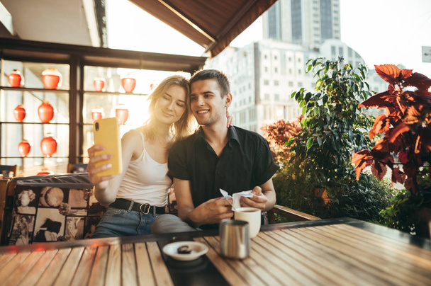Feliz casal amoroso fazendo selfie no café à mesa no fundo do pôr do sol. Bonito casal sorrindo sentado em aconchegante. restaurante no terraço e tirar selfie posando na câmera do smartphone
. - Foto, Imagem