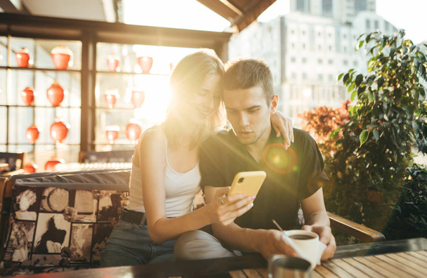 Młody mężczyzna i dziewczyna siedzący na tarasie w restauracji i za pomocą smartfona na tle zachodu słońca, dziewczyna uśmiechnięta, facet wyglądający zaskoczony twarz na ekranie smartfona. Randka w przytulnej kawiarni koncepcji. - Zdjęcie, obraz