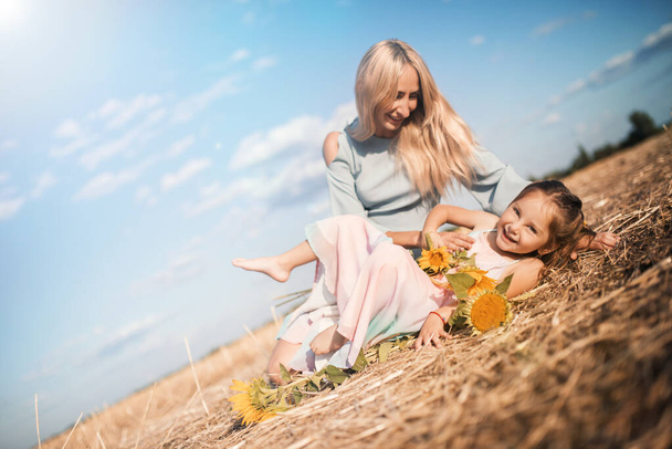 Charming young mother is having fun with her little daughter sitting on a field with sunflowers on a warm sunny summer day. Weekend vacation concept with family - Photo, image