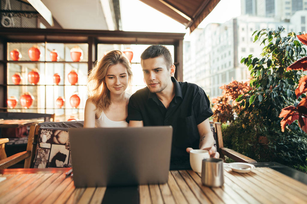 Knappe jongeman en meisje zitten op het terras in een gezellig café en gebruiken een laptop, gericht op het scherm. Jonge paar studenten studeren in cafe op terras. - Foto, afbeelding