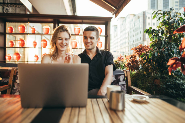 Happy couple of students sitting with laptop in cafe and sunset, looking at screen and smiling. Beautiful young man and girl watching funny video on internet at table in cafe and rejoicing. - 写真・画像