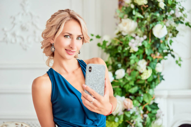 Attractive young woman with a stylish hairstyle in an elegant dress makes selfie posing in the style of a modern studio. The concept of the beauty of luxury and gadgets - Fotografie, Obrázek