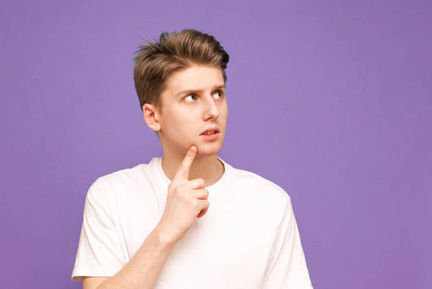 Portrait of a guy in a white T-shirt stands on a purple background, looks aside and thinks. Thoughtful young man poses on a blue background and looks at a place for advertising. Isolated. - Zdjęcie, obraz