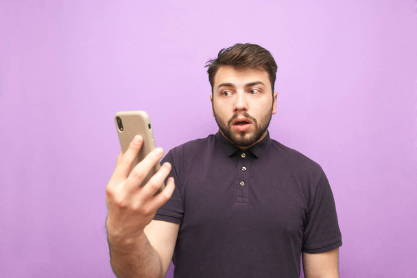 Homme étonnant avec une barbe se tient avec un smartphone dans ses mains et semble confondu avec un visage confus. Homme drôle tient un smartphone dans ses mains, isolé sur un fond violet
 - Photo, image
