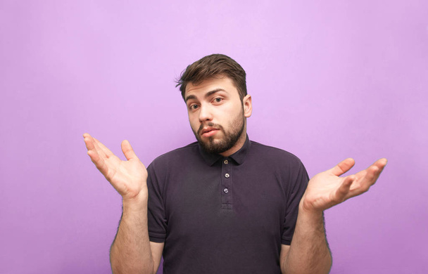 El hombre barbudo divertido con una camiseta oscura muestra una confusión sobre un fondo púrpura, mira a la cámara y hace una cara desconcertada. Muestra gestos que no saben nada. Aislado
 - Foto, Imagen