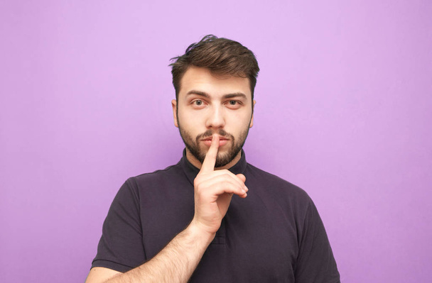 Man in een donker T-shirt op een paarse achtergrond houdt zijn vinger om zijn mond en kijkt bedachtzaam naar de camera.Gericht man is geïsoleerde paarse achtergrond, een portretje.Man toont een teken van stilte - Foto, afbeelding