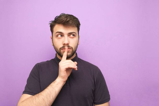 Portrait of an adult man with a beard thoughtfully looking sideways to a blank place and showing a sign of silence holding a finger near his mouth on a purple background. Isolated. - 写真・画像