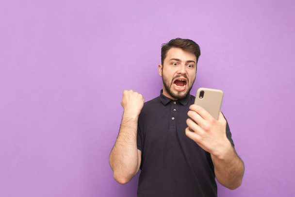 Positive young man holds a smartphone in his hands, looks at the screen and rejoices with his hands raised. Guy smiles and rejoices in success isolated on a purple background. Copyspace - Photo, Image