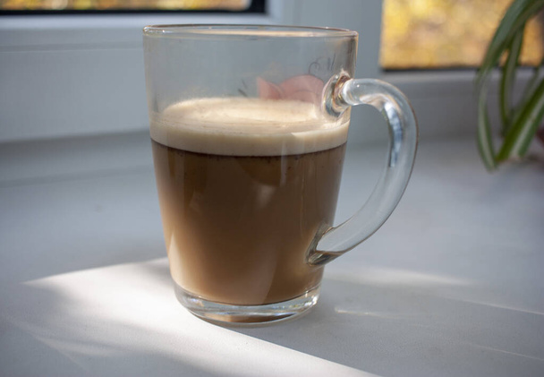 Ízletes reggeli kávé tejjel egy üvegbögrében dupla falakkal az ablakpárkányon a napsütésben. - Fotó, kép