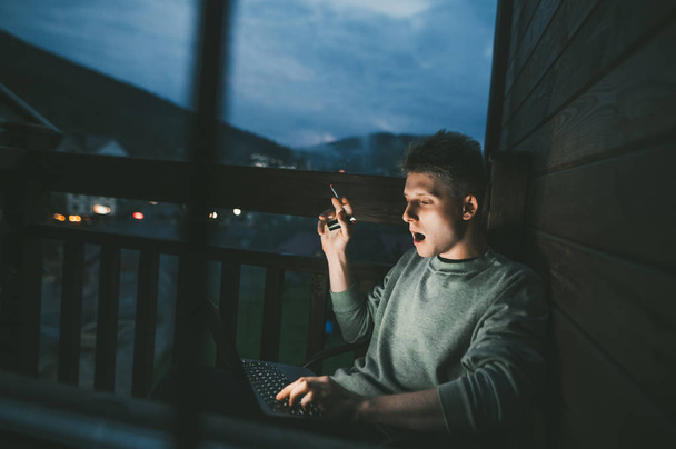 Der junge Mann raucht auf dem Balkon eine Zigarette und benutzt abends in einem Landhaus einen Laptop, schaut in den Bildschirm. Mann sitzt mit Zigarette auf dem Balkon in einem Stuhl und arbeitet an einem Laptop - Foto, Bild