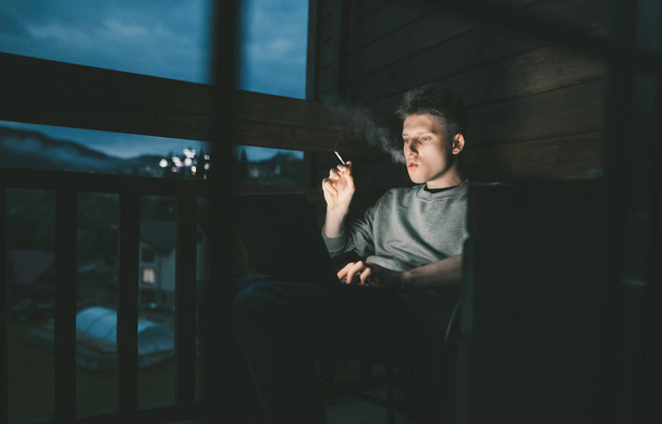 junger Mann raucht eine Zigarette und arbeitet am Abend an einem Laptop auf einem Wohnbalkon.Freelancer Kerl arbeitet an einem Laptop spät in der Nacht und atmet Zigarettenrauch aus, arbeitet in einem Landhaus - Foto, Bild