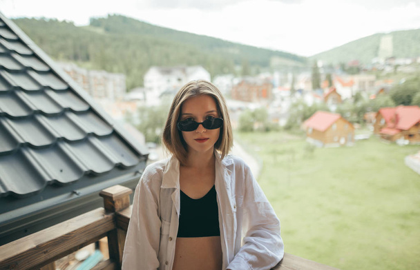 Mulher atraente em roupas casuais elegantes e óculos de sol posando na varanda em uma casa de campo, olhando para a câmera com um rosto sério no fundo das montanhas e casas paisagem
 - Foto, Imagem