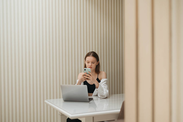 Piękna dziewczyna biznesowa używa smartfona i laptopa przy stole w domu przy stole w mieszkaniu. Atrakcyjna kobieta w luźnych ciuchach, pracująca w domu z laptopem, portret w pokoju. Wolny strzelec - Zdjęcie, obraz