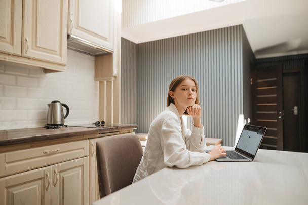 Portrait d'une étudiante assise à un bureau dans la cuisine et utilisant un ordinateur portable, avec un visage sérieux regarde à la caméra. Belle fille travaillant sur ordinateur portable à la table à la maison dans l'appartement
 - Photo, image