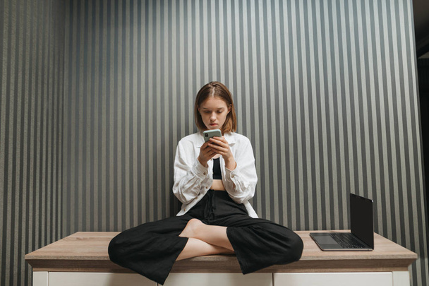 Beautiful girl in casual clothes sits on a bench indoors on a background of gray wall with wallpaper, uses a phone, a laptop stands near her.Freelancer girl is resting with a smartphone in her hands - Photo, Image