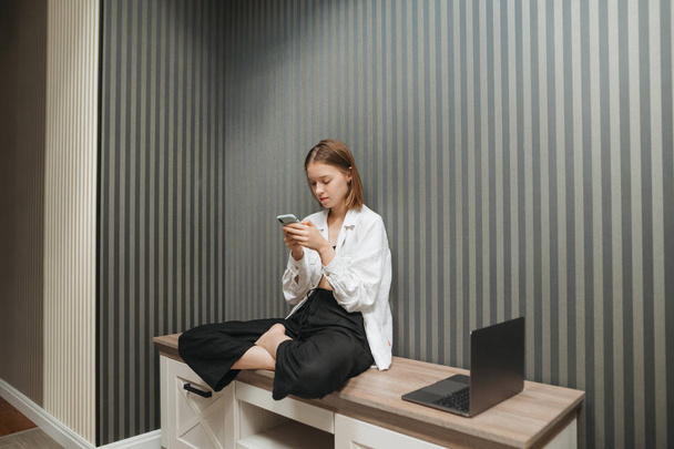 Pretty girl in stylish clothes sits on a bedside table at home in the apartment and uses a smartphone, gray wall background, the laptop stands on the side. Freelancer girl's portrait works at home - Photo, Image